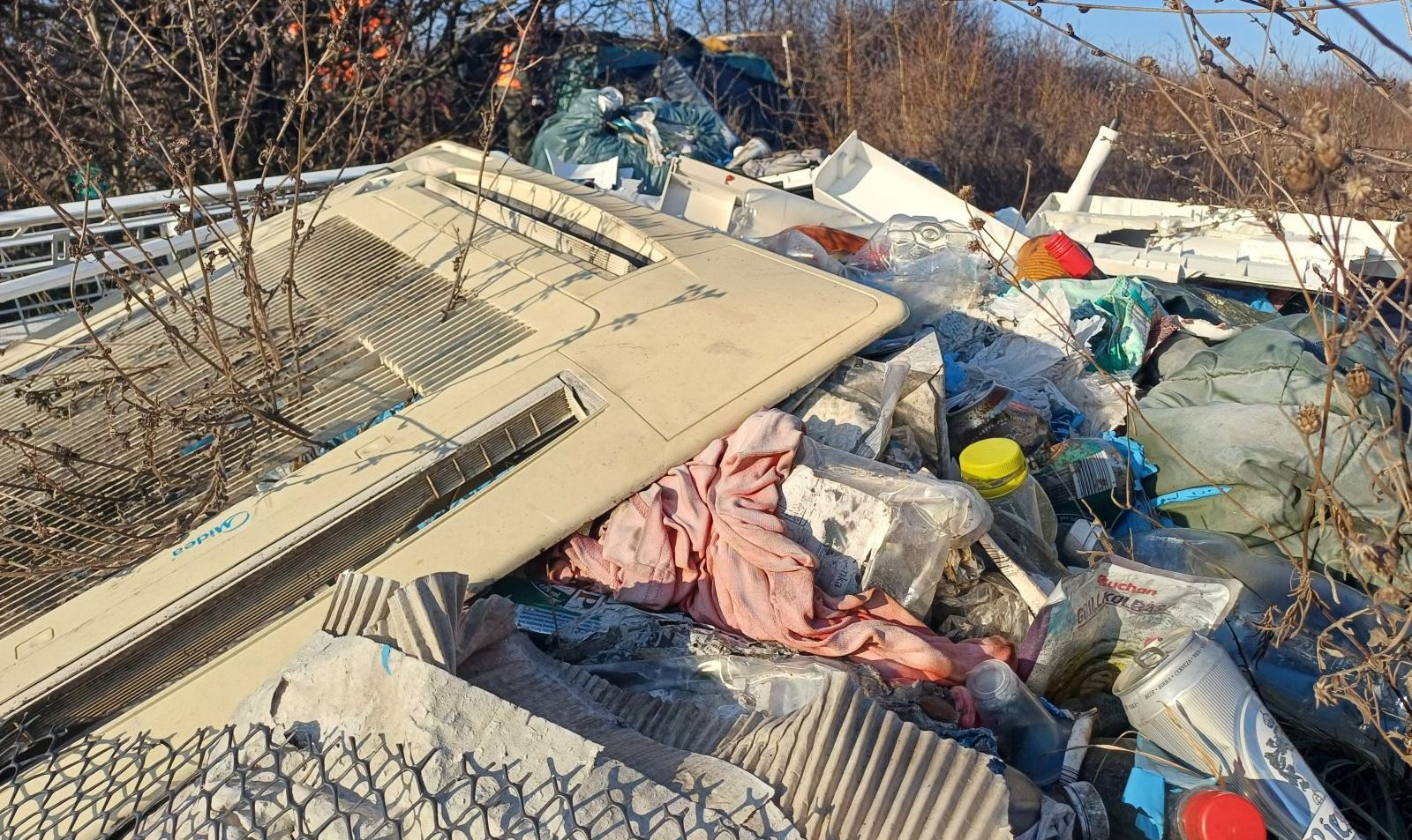 A természetbe kidobott szeméthalom van a képen. A szemét között van többek között ajtó, ruha, italospalackok